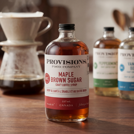 Bottle of Provisions Maple Brown Sugar syrup on a wooden surface with a coffee setup in the background.