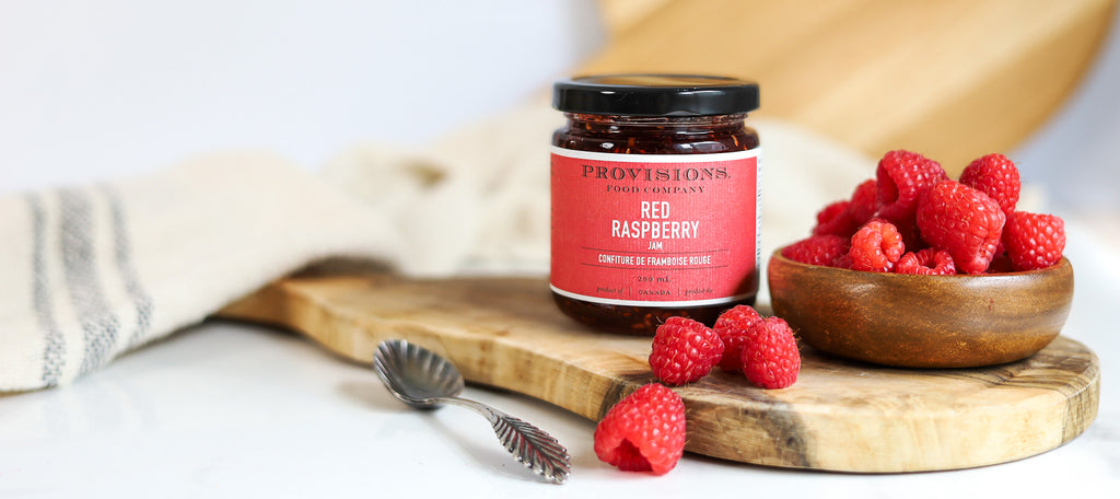 Provisions Red Raspberry Jam beside juicy red raspberries sitting in a small wood bowl